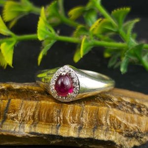 Könnte beinhalten: Ein silberner Ring mit einem tropfenförmigen, tiefroten Edelstein, umgeben von kleinen, klaren Steinen. Der Ring liegt auf einer strukturierten, braunen Oberfläche, mit grünem Laub im Hintergrund. Der Ring hat ein glattes, poliertes Band.