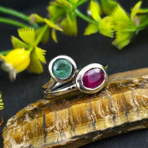 Könnte beinhalten: Ein silberner Ring mit zwei ovalen Edelsteinen: einem grünen Smaragd und einem roten Rubin. Der Ring liegt auf einer strukturierten, braunen Oberfläche, mit grünem und gelbem Laub im Hintergrund. Die Edelsteine sind in einer Fassung gefasst.