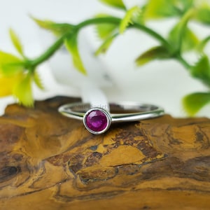 Könnte beinhalten: Ein silberner Ring mit einem runden, leuchtend roten Edelstein. Der Ring liegt auf einer Holzoberfläche mit grünem und gelbem Laub im Hintergrund. Der Edelstein ist der Blickfang des Rings.