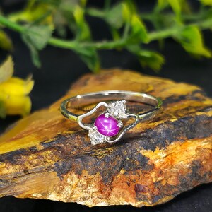 Könnte beinhalten: Ein silberner Ring mit einem sternförmigen, pinkfarbenen Edelstein in der Mitte, akzentuiert durch kleinere, klare Steine. Der Ring liegt auf einer strukturierten, braun-gelben Oberfläche, mit grünem Laub im Hintergrund.