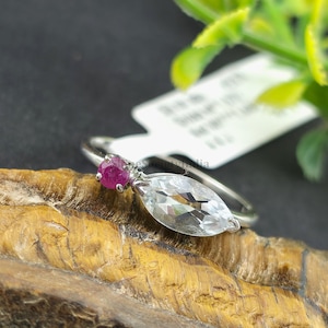 Könnte beinhalten: Ein silberner Ring mit einem kleinen, runden, roten Edelstein und einem größeren, marquiseförmigen, klaren Edelstein. Der Ring liegt auf einer natürlichen Holzoberfläche mit grünem Laub im Hintergrund.