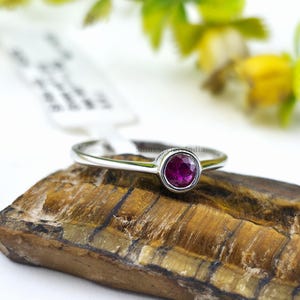 Könnte beinhalten: Ein silberner Ring mit einem runden, tiefroten Edelstein. Der Ring liegt auf einer strukturierten, braunen Oberfläche, mit einem verschwommenen Hintergrund aus grünem und gelbem Laub. Der Edelstein ist der Mittelpunkt des Rings.