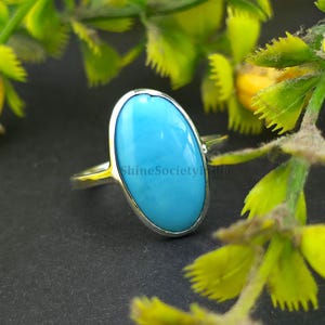 Könnte beinhalten: Ein Silberring mit einem großen, ovalen Türkis-Edelstein. Der Stein ist in einer Silberfassung gefasst, und das Band ist schlichtes Silber. Der Ring wird vor einem dunklen Hintergrund mit grünem und gelbem Laub gezeigt. Der Text "ShineSocietyIndia" ist sichtbar.
