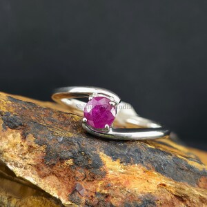 Könnte beinhalten: Ein silberner Ring mit einem runden, tiefrosa Rubin-Edelstein. Das Band des Rings umschließt den Stein und erzeugt ein modernes, elegantes Design. Der Ring wird auf einer strukturierten, braunen Oberfläche vor dunklem Hintergrund präsentiert.