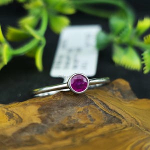 Könnte beinhalten: Ein silberner Ring mit einem runden, tiefrosa Edelstein. Der Ring liegt auf einer braunen Oberfläche, mit grünem Laub im Hintergrund. Der Edelstein ist der Mittelpunkt des Rings.