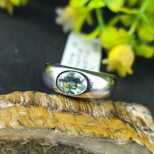 Puede incluir: Un anillo de plata con una piedra preciosa ovalada de color verde claro. El anillo tiene una banda ancha y un acabado pulido. La piedra preciosa está engastada en un bisel. El anillo se exhibe sobre un trozo de madera, con un fondo borroso de flores amarillas.