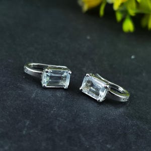 Könnte beinhalten: Ein Paar silberfarbene Ohrringe mit rechteckigen, klaren Edelsteinen. Die Ohrringe sind auf einem dunklen Untergrund platziert, mit einem verschwommenen grünen und gelben Hintergrund. Die Edelsteine sind facettiert.