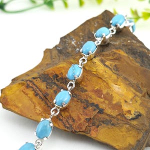 Könnte beinhalten: Ein silbernes Armband mit ovalen, hellblauen Türkissteinen. Die Steine sind in einzelnen Silberfassungen gefasst und miteinander verbunden. Das Armband wird auf einer braunen und beigen Steinoberfläche präsentiert.