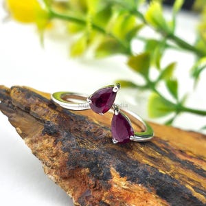Könnte beinhalten: Ein silberner Ring mit zwei tropfenförmigen, tiefroten Edelsteinen. Das Design des Rings weist die Edelsteine in entgegengesetzte Richtungen auf. Der Ring wird auf einem Holzstück mit verschwommenem grünem Hintergrund präsentiert.
