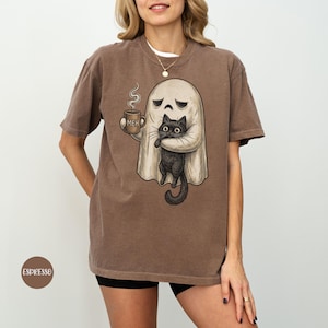 May include: A brown short-sleeve t-shirt featuring a graphic of a ghost holding a steaming mug labelled MEH and hugging a black cat. The ghost has a sad expression, and the cat has large eyes. The shirt has a crew neckline and a relaxed fit.