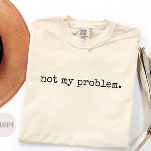 May include: An ivory-colored short-sleeve t-shirt featuring the black text "not my problem." printed across the chest. The shirt has a crew neckline and a Comfort Colors brand label visible on the inside of the collar.