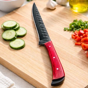 Könnte beinhalten: Ein schwarzes und rotes Küchenmesser mit strukturierter Klinge liegt auf einem Holzschneidebrett. Auf dem Brett befinden sich auch geschnittene Gurken, Tomaten, Knoblauch und Öl, was auf die Zubereitung von Speisen hindeutet. Das Messer hat einen roten Griff mit weißen Punkten.