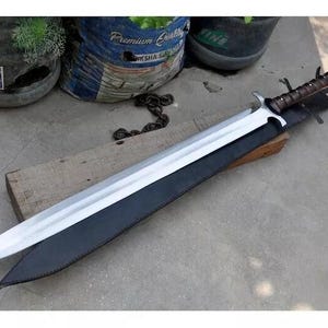 May include: A long, silver sword with a brown handle and a black sheath. The blade is sharp and reflective, and the handle is wrapped in a dark brown material. The sword is resting on a wooden block.