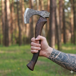 May include: A hand-held axe with a dark wooden handle and a silver-colored blade. The axe has a curved handle and a textured grip. The background is a blurred forest scene.