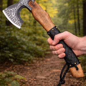 Könnte beinhalten: Eine Axt im Wikinger-Stil mit einem Holzgriff und einem Metallkopf mit aufwendigen Gravuren. Der Griff ist mit schwarzem Leder mit Messingnieten umwickelt. Eine schwarze Lederschnur ist am unteren Ende des Griffs befestigt. Die Axt wird vor einem verschwommenen Waldboden gehalten.