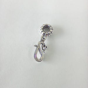 May include: A silver-coloured metal charm bead featuring a dangling snake pendant. The bead has a circular, textured top loop for attachment, while the snake is depicted in a coiled position with a smooth, polished finish and small dark eyes.