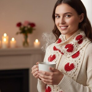 Puede incluir: Una mujer con un suéter color crema y una bufanda de ganchillo con diseños de corazones rojos. Sostiene una taza blanca, de la que sale vapor. El fondo incluye una chimenea y velas encendidas.