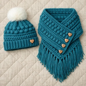 May include: A teal crochet hat with a white pom-pom and a matching scarf with fringe. The hat and scarf set feature a textured stitch pattern and wooden heart-shaped buttons. A cozy winter accessory.