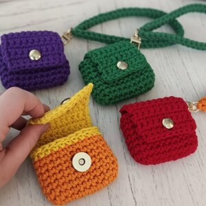 May include: A collection of small, crocheted pouches in various colors: purple, green, red, and orange. One pouch is open, revealing a yellow interior. Each pouch has a flap secured with a silver snap. A green crocheted strap is attached to one of the pouches.