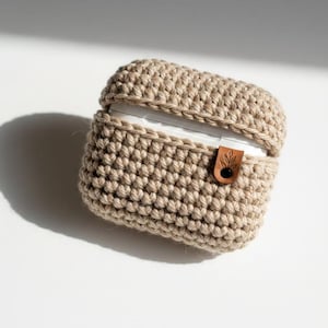 Könnte beinhalten: Eine beige Häkelhülle für kabellose Ohrhörer. Die Hülle besteht aus dickem, strukturiertem Garn und hat einen kleinen, braunen Lederakzent mit einem schwarzen Knopfverschluss. Die Oberseite der Hülle ist offen und gibt die weißen Ohrhörer frei.