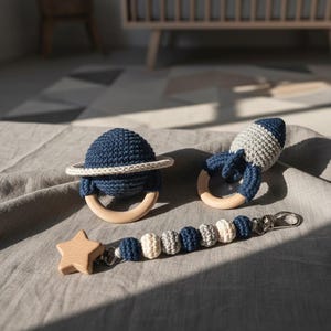 Puede incluir: Un conjunto de tres juguetes para bebés: un sonajero azul marino en forma de planeta con un anillo blanco, un sonajero en forma de cohete gris y azul marino, y un clip para chupete con una estrella de madera y un diseño de cuentas. Los juguetes son de ganchillo y madera.