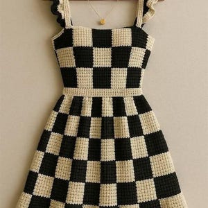 May include: A handmade, black and cream-colored crochet dress with a checkerboard pattern. The dress features ruffled straps and a square neckline. A gold necklace with a small pendant hangs on a hanger behind the dress.