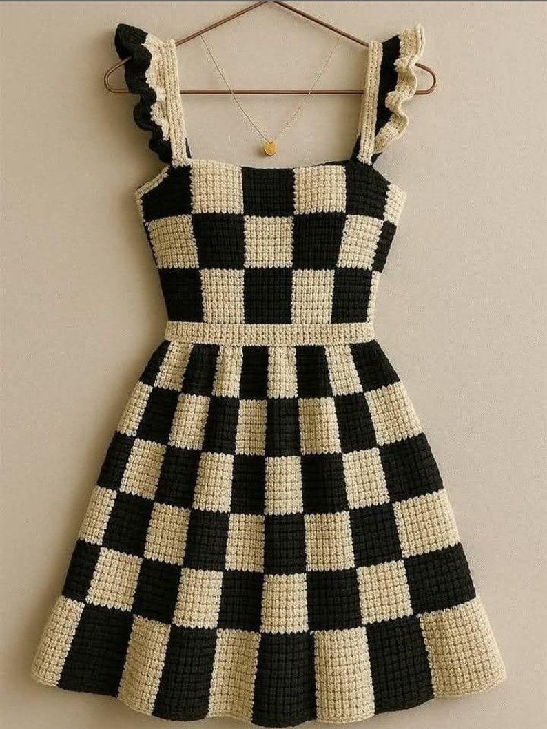 May include: A handmade, black and cream-colored crochet dress with a checkerboard pattern. The dress features ruffled straps and a square neckline. A gold necklace with a small pendant hangs on a hanger behind the dress.