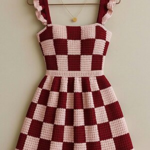 May include: A handmade crochet dress in a checkerboard pattern of burgundy and light pink. The dress features ruffled straps and a fitted bodice that flares into a skirt. A gold heart pendant hangs from a delicate chain.