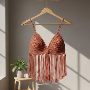 Può includere: Un top all'uncinetto color terracotta con scollo a V profondo e frange. Il top è appeso a una gruccia di legno, mostrando la sua consistenza e il suo design fatti a mano. Le frange pendono sotto il busto, aggiungendo un tocco bohémien.
