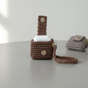 May include: Crocheted brown case for wireless earbuds with a matching strap and a snap closure. A second, smaller crocheted case in a light brown color is also present, with a strap and snap closure. The cases are on a light gray surface.