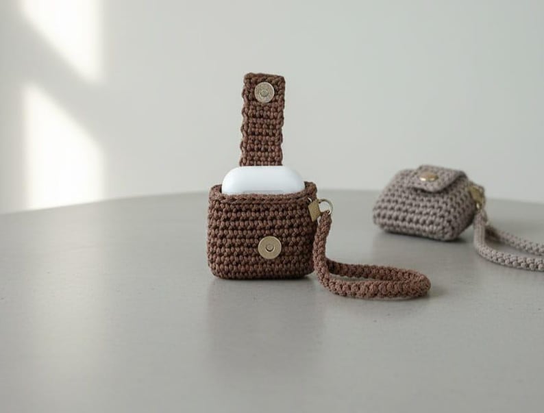 May include: Crocheted brown case for wireless earbuds with a matching strap and a snap closure. A second, smaller crocheted case in a light brown color is also present, with a strap and snap closure. The cases are on a light gray surface.