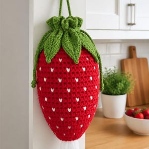 Può includere: Una borsa a forma di fragola rossa all'uncinetto con accenti bianchi a forma di cuore e foglie verdi. La borsa è appesa a una parete bianca. Un cordino verde viene utilizzato per appendere la borsa. La borsa è progettata per contenere sacchetti di plastica.