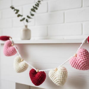 May include: A crochet heart garland in red, pink, and cream. The hearts are strung on a white cord with wooden beads. The garland is displayed against a white brick wall, creating a charming, romantic aesthetic. Ideal for Valentine's Day.