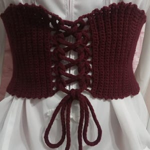 May include: A burgundy crochet corset with a lace-up front. The corset has a scalloped edge and is shown over a white garment. The crochet work is detailed, with a textured appearance. The corset is a top, likely for fashion or costume.