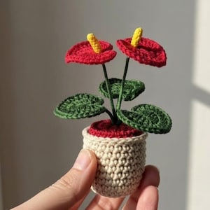May include: A handmade crochet plant featuring two red flowers with yellow centers, green leaves, and a beige pot with a red rim. The miniature plant is held in a hand, showcasing the intricate details of the craft.