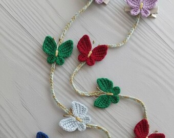 May include: A collection of colorful, crocheted butterfly hair clips. The butterflies are in various colors, including purple, green, red, blue, white, and yellow. Each butterfly is attached to a clip with a braided cord.