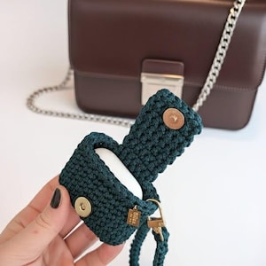Può includere: Una custodia per AirPods all'uncinetto verde scuro con una chiusura color oro e un cinturino da polso abbinato. La custodia è aperta, rivelando gli AirPods bianchi all'interno. Una borsa in pelle marrone con una catena d'argento è sullo sfondo.