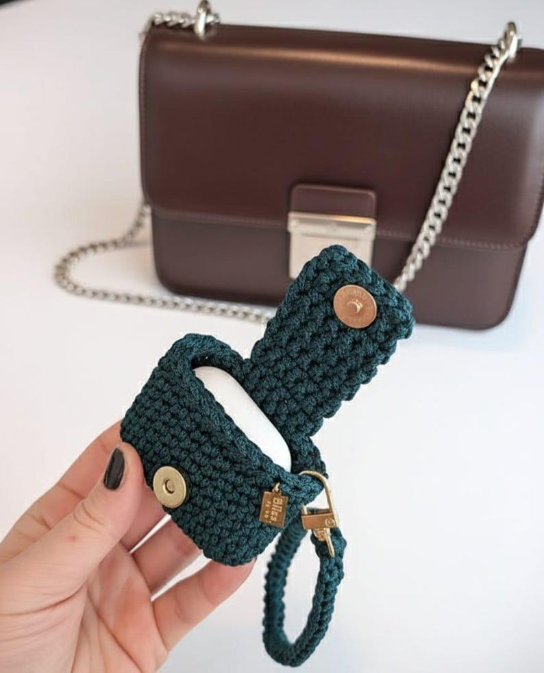 May include: A dark teal crocheted AirPods case with a gold-colored clasp and a matching wrist strap. The case is open, revealing white AirPods inside. A brown leather handbag with a silver chain strap is in the background.