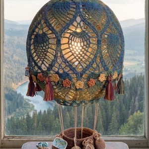 May include: A handcrafted, crochet hot air balloon lamp with a blue and green patterned design. The lamp features decorative tassels and a woven basket base. Additional decorative items include a globe, a ring, and a journal.