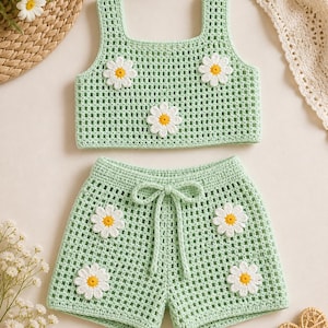 May include: A light green crochet two-piece set comprising a crop top and shorts, both decorated with daisy appliqués. The top has a square neckline, and the shorts have a drawstring waist. The daisy flowers have white petals and yellow centres.