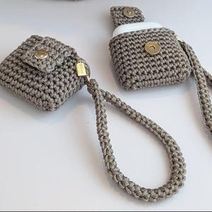 May include: Two crocheted cases in a neutral gray color. One case is square with a flap and gold button closure. The other case is designed for earbuds and has a matching strap. Both cases have a gold tag attached.