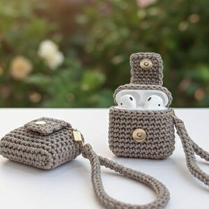 May include: Two crocheted AirPod cases in a neutral gray color. One case is open, revealing white earbuds, and the other is closed. Both cases have a button closure and a matching wrist strap. The background is blurred.