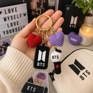 May include: A gold-toned metal keychain featuring four crocheted charms: a red heart, a purple heart, a black rectangular tag with the white text BTS and a logo, and a white and black spherical charm with the text BTS and a small red accent.