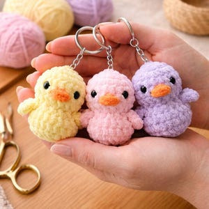 May include: Three handmade crochet chick keychains in yellow, pink, and lavender. Each chick has a small orange beak and black eyes. The keychains are held in a person's hand, with yarn and scissors in the background.