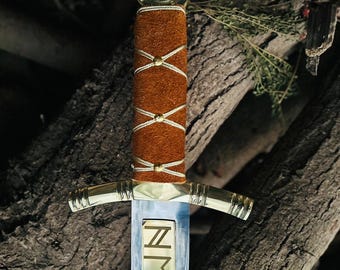 May include: A decorative sword with a brown suede handle wrapped with white and gold cord. The blade is silver with gold lettering that reads "NMEMMA". The sword has a gold crossguard and a circular gold pommel.