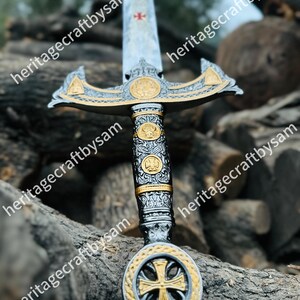 May include: An ornamental sword with a silver blade and gold detailing. The hilt is intricately designed with a cross motif. The sword is set against a backdrop of wooden logs, highlighting its craftsmanship and historical appeal.