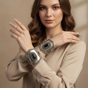 Può includere: Due braccialetti a polsino argentati. Ogni braccialetto presenta una pietra nera ovale incastonata in una cornice decorativa. Le cornici hanno un design ornato di ispirazione vintage con piccoli accenti rotondi. I braccialetti sono indossati ai polsi.