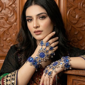 Könnte beinhalten: Eine Frau trägt aufwendigen blauen und silbernen Handschmuck. Das Set umfasst ein Armband, eine Handkette und einen Ring, alle mit blauen Edelsteinen und baumelnden silbernen Akzenten. Sie trägt ein schwarzes Oberteil mit farbenfrohen Stickereien.
