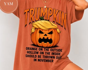 Peut inclure: Un t-shirt couleur rouille avec un motif de citrouille d'Halloween aux cheveux blonds. Le texte indique TRUMPKIN en haut et ORANGE ON THE OUTSIDE HOLLOW ON THE INSIDE SHOULD BE THROWN OUT IN NOVEMBER en bas. Une étiquette circulaire YAM est visible.