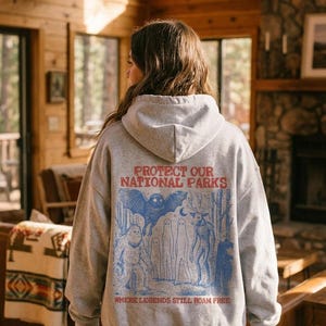 May include: A light gray hooded sweatshirt featuring a red and blue graphic on the back. The text reads PROTECT OUR NATIONAL PARKS and WHERE LEGENDS STILL ROAM FREE. The illustration depicts a bat, a sasquatch, ghosts, and a deer-like creature.